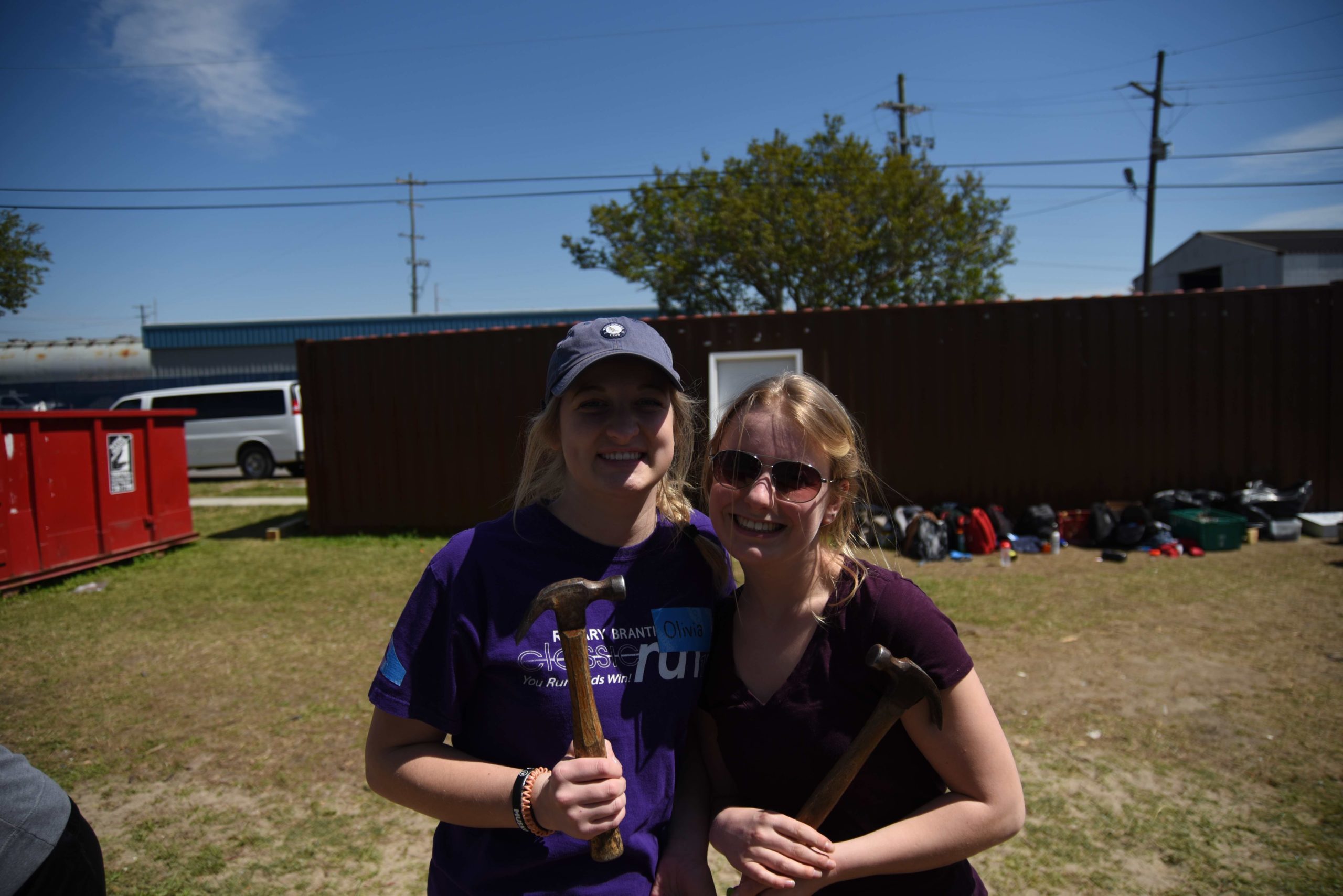 Two people wearing purple t-shirts smile at the camera while holding hammers