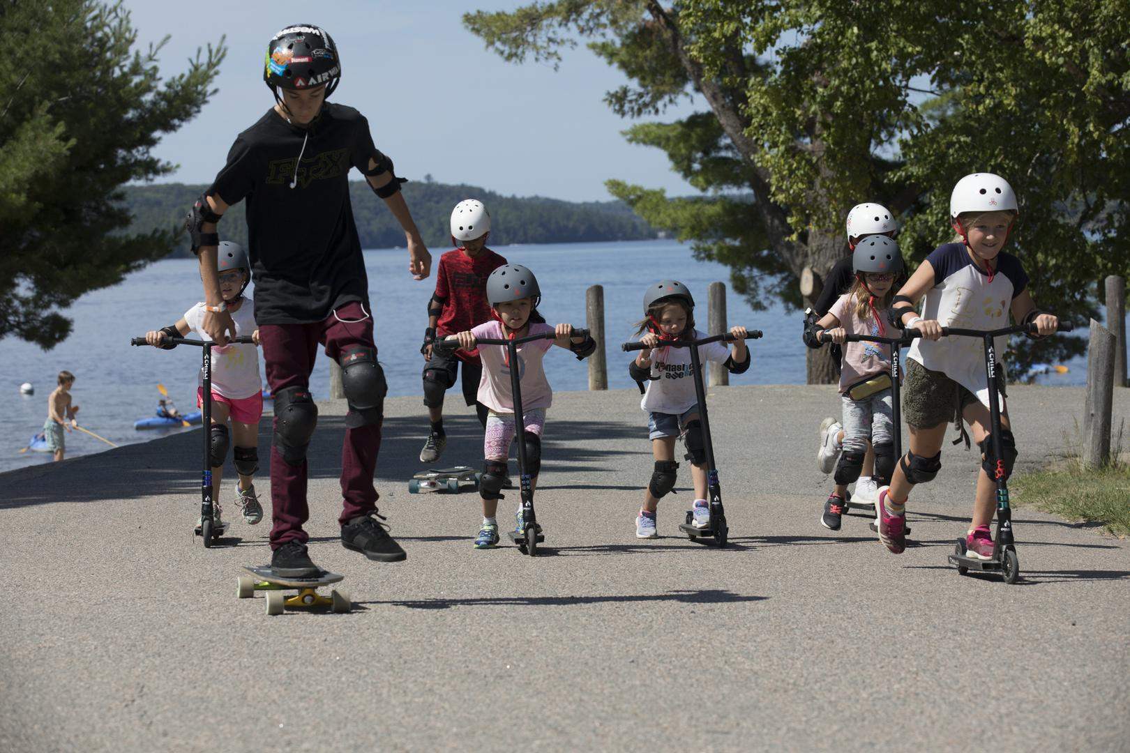 young boys and girls use scooters in front of a lake